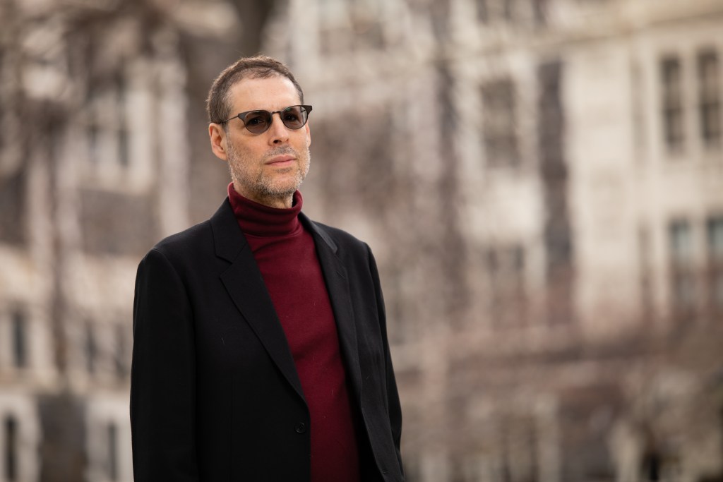 Ray Gallon wearing sunglasses and a red polo neck sweater with black jacket outside with city buildings in the backdrop