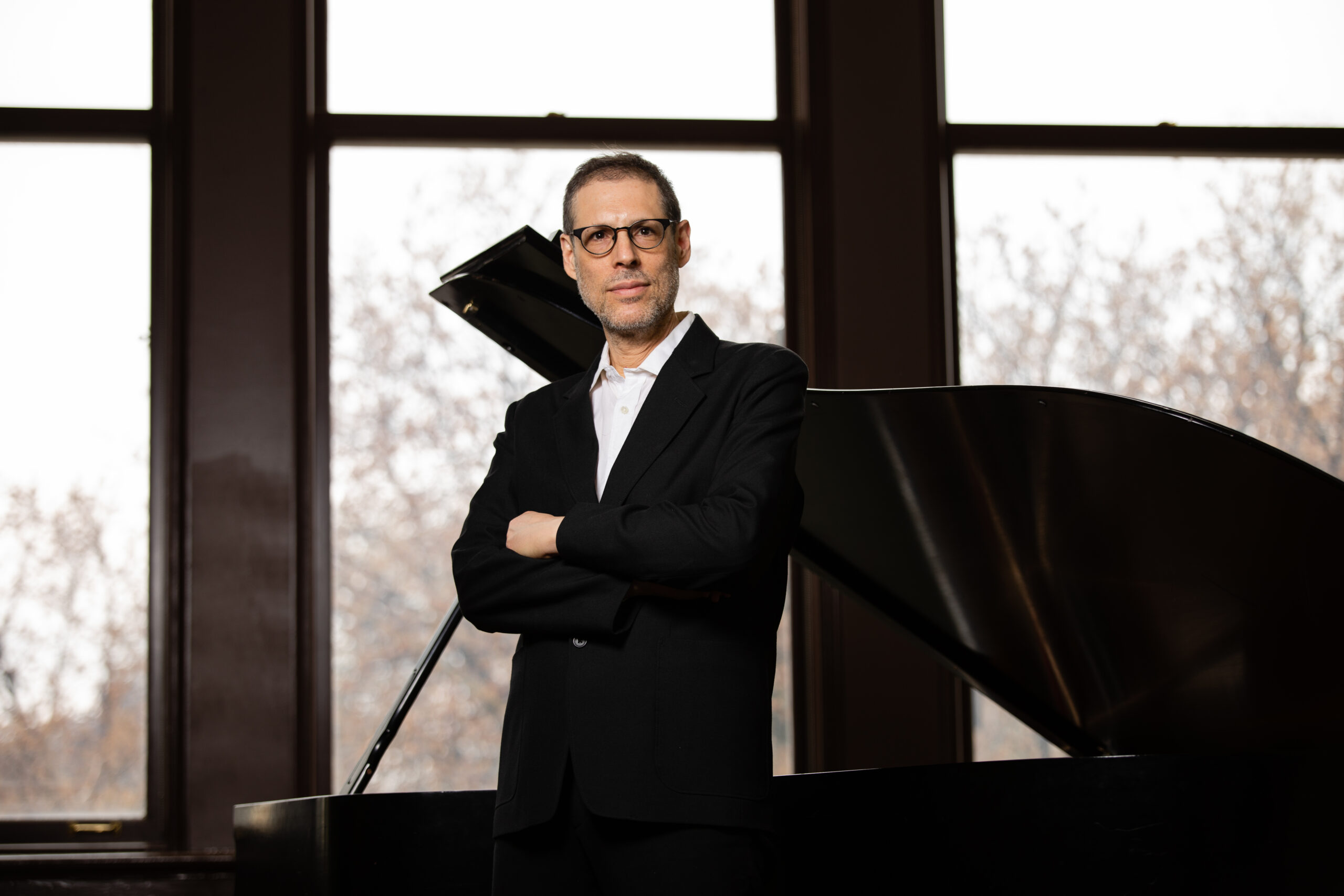Ray Gallon wearing a black jacket and white shirt standing in front of a grand piano by a large window